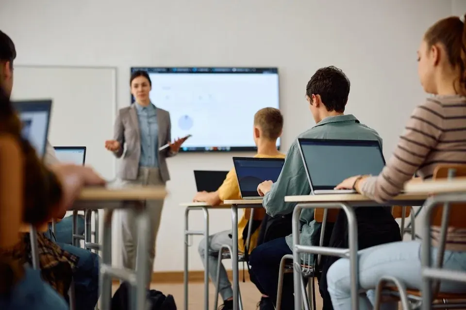  Schüler sitzen mit Laptops in Reihen, Lehrerin vorne präsentiert auf Bildschirm — Blick in den Klassenraum während des Unterrichts.