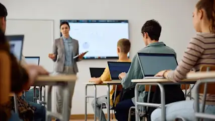 Schüler sitzen mit Laptops in Reihen, Lehrerin vorne präsentiert auf Bildschirm — Blick in den Klassenraum während des Unterrichts.