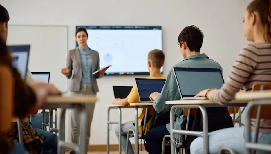 Schüler sitzen mit Laptops in Reihen, Lehrerin vorne präsentiert auf Bildschirm — Blick in den Klassenraum während des Unterrichts.
