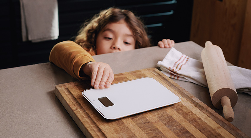 Auf einer Arbeitsplatte liegen ein Schneidebrett, eine Waage und eine Teigrolle. Ein Geschirrtuch ist im Hintergrund zu sehen.