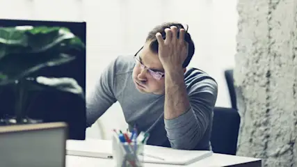 Ein Mann mit Brille, in grauem Pullover, sitzt am Schreibtisch, die Hand am Kopf. Vor ihm Stifte, Notizbuch, Laptop. Links ein Monitor mit Pflanze, rechts eine Betonwand.