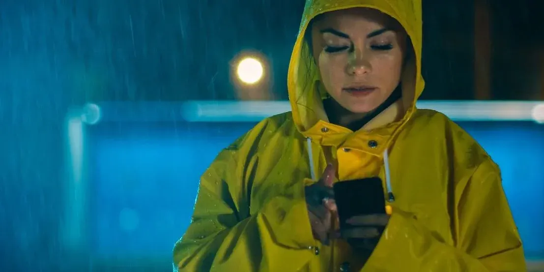 Eine Frau im gelbem Regenmantel steht im strömenden Regen und bedient ihr Smartphone.