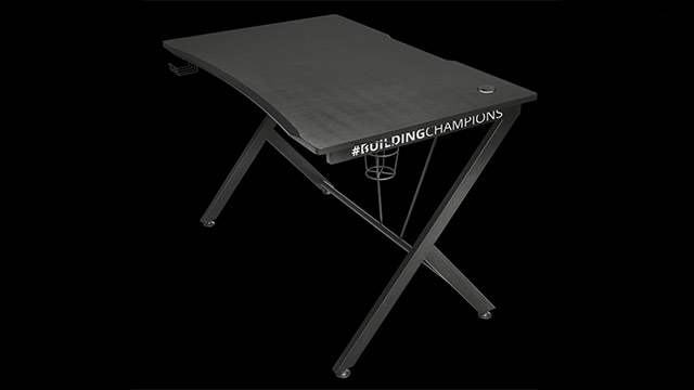 Schwarzer Schreibtisch mit gekreuzten Beinen und Getränkehalter vor schwarzem Hintergrund. #BUILDINGCHAMPIONS ist auf der Seite gedruckt.