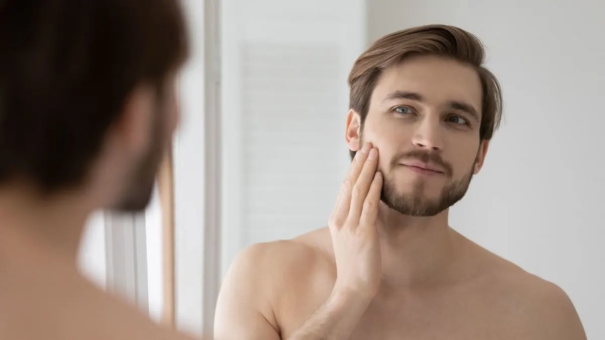 Ein lächelnder Mann betrachtet sich im Spiegel und berührt seinen Bart.