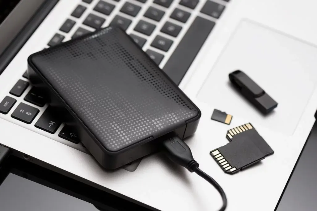 Externes schwarzes USB‑Festplattenlaufwerk auf Laptop‑Tastatur, daneben SD‑Karten und USB‑Stick, Kabel angeschlossen.