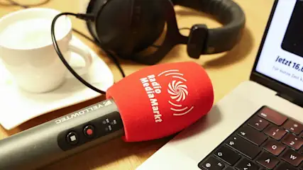 Graues Mikrofon mit rotem "Radio MediaMarkt" Schaumstoff, ruht auf einem silbernen Laptop. Im Hintergrund weiße Tasse mit Getränk und Kopfhörer.