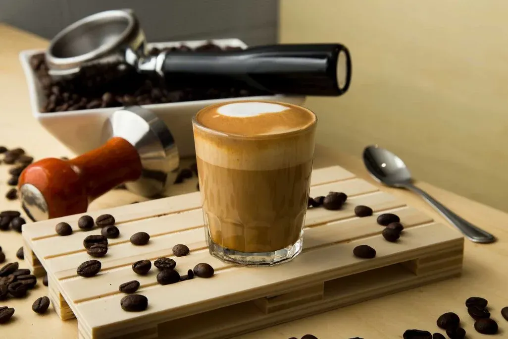 Kaffeeglas mit weißem Schaum auf heller Holzpalette, Kaffeebohnen verstreut. Tamper, Siebträger und Löffel im Hintergrund.
