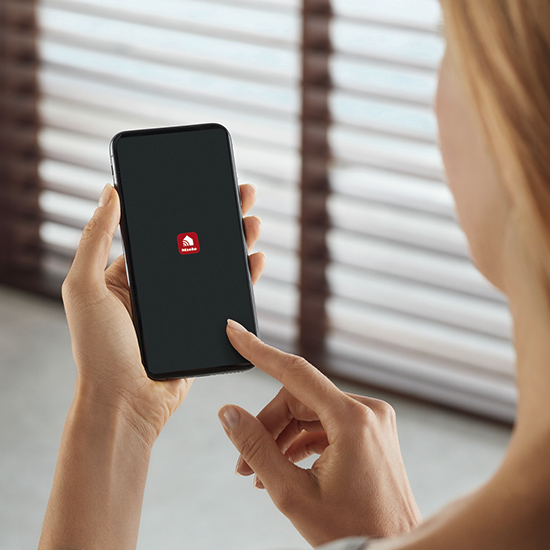 Eine Person hält ein schwarzes Smartphone mit einem roten Logo vor einem Hintergrund mit Jalousien.