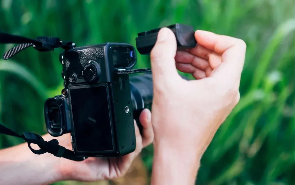 Hände halten eine schwarze Kamera mit offenem Batteriefach. Eine Hand führt einen schwarzen Akku in das Fach ein. Unscharfes grünes Laub im Hintergrund.
