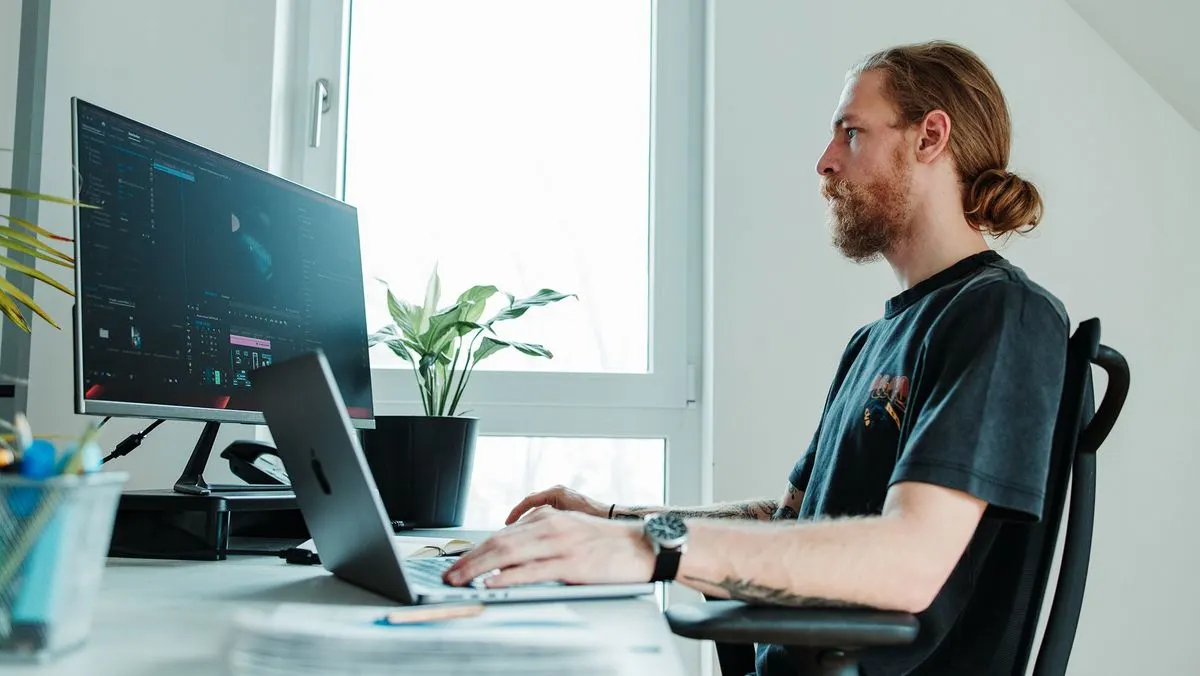 Person tippt am Schreibtisch auf einem Laptop vor einem großen Monitor mit Software. Im Hintergrund: helles Fenster, grüne Pflanze.
