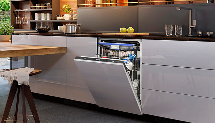 Vollintegrierter KOENIC Einbau-Geschirrspüler mit geöffneter Tür und blauer  LED-Innenbeleuchtung. Ober- und Unterkorb sind mit Tellern, Gläsern und Tassen  beladen. Der Geschirrspüler ist in eine moderne Küche mit hochglänzenden  weiß-grauen Fronten und dunkler Arbeitsplatte eingebaut. Im Hintergrund  offene Regale mit Küchenutensilien, links ein Holztresen.
