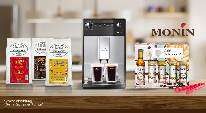 Kaffeemaschine mit Bohnen, Sirup und Zubehör auf einem Tisch. Im Hintergrund ist eine Küche erkennbar.