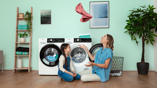 Eine Frau und ein Kind sitzen vor Waschmaschinen, während ein rosafarbenes Kleidungsstück in der Luft schwebt.