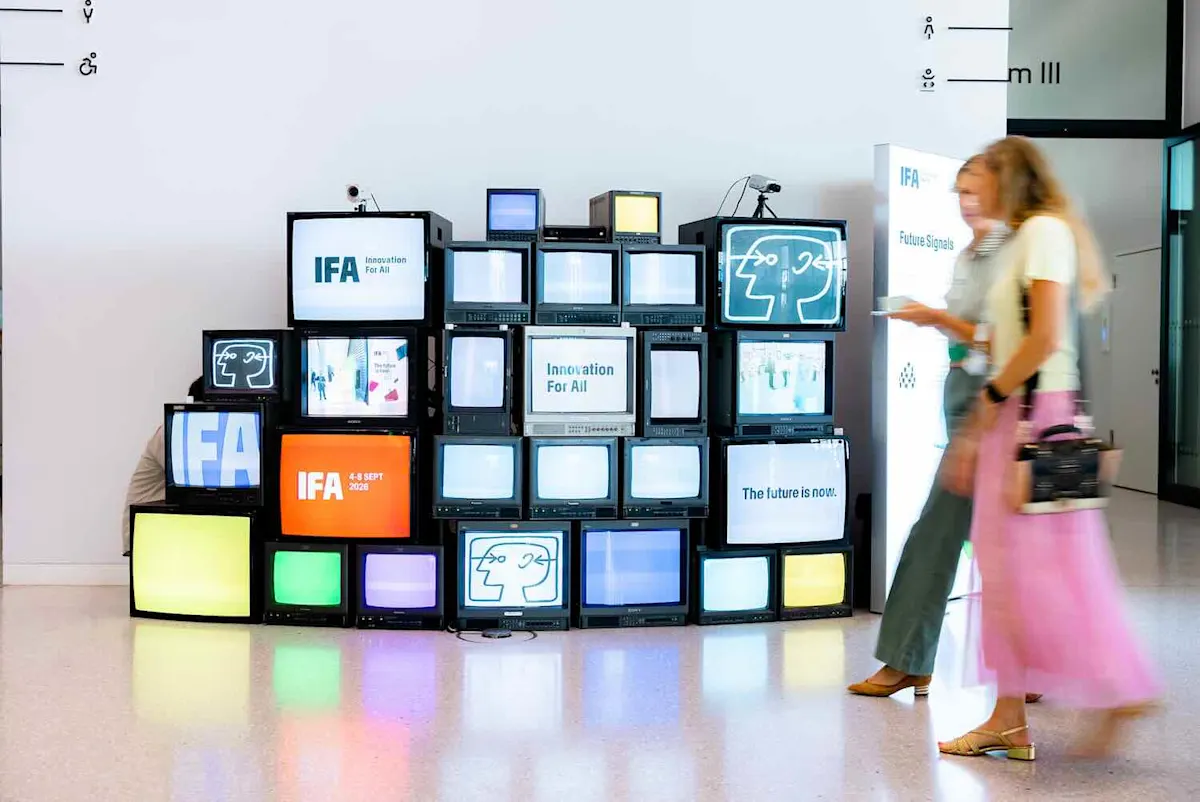 Zwei Frauen gehen durch eine IFA-Messehalle. Im Hintergrund sind Fernseher übereinander gestapelt, die das IFA-Logo und IFA-Slogans zeigen.