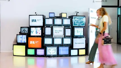 Zwei Frauen gehen durch eine IFA-Messehalle. Im Hintergrund sind Fernseher übereinander gestapelt, die das IFA-Logo und IFA-Slogans zeigen.