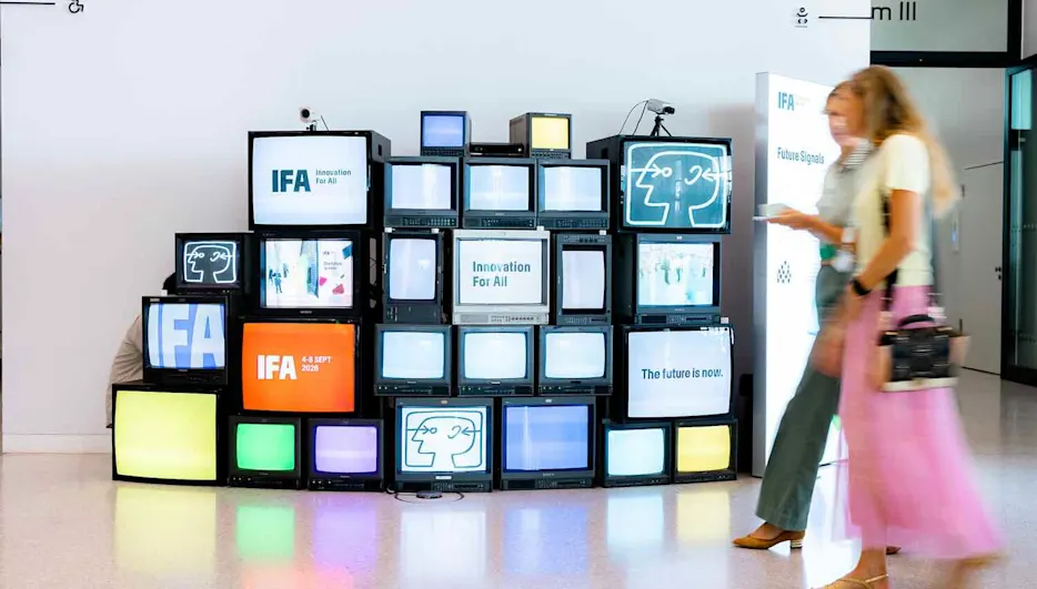 Zwei Frauen gehen durch eine IFA-Messehalle. Im Hintergrund sind Fernseher übereinander gestapelt, die das IFA-Logo und IFA-Slogans zeigen.