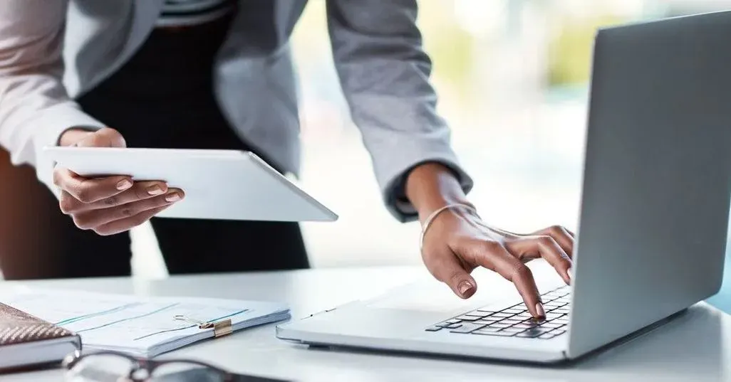 Hände am Tisch halten ein Blatt Papier und tippen gleichzeitig auf dem Laptop.