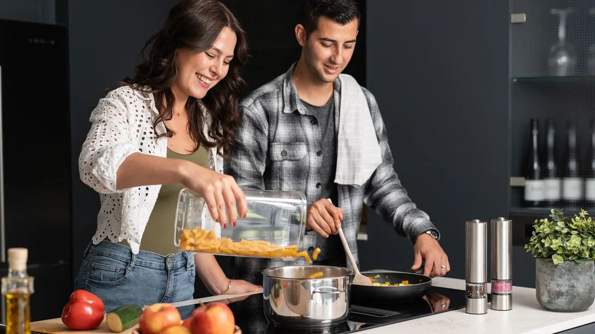 Zwei Personen kochen gemeinsam in einer modernen Küche; eine Frau schüttet Nudeln in einen Topf, ein Mann rührt in einer Pfanne