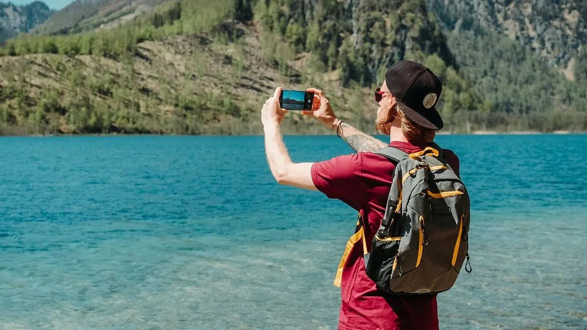 Person mit Rucksack fotografiert Landschaft an einem See mit Smartphone.