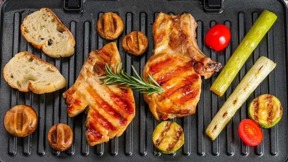 Zwei gegrillte Schweinekoteletts mit Rosmarin, Brotscheiben, Pilzen, Tomaten, Lauch und Zucchini auf einer Grillpfanne.