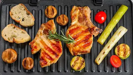 Zwei gegrillte Schweinekoteletts mit Rosmarin, Brotscheiben, Pilzen, Tomaten, Lauch und Zucchini auf einer Grillpfanne.