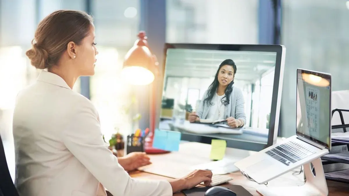 Frau in heller Jacke tippt am Schreibtisch, sieht Videoanruf einer anderen Frau auf Monitor. Ein Laptop auf Ständer und eine rote Lampe sind dort.