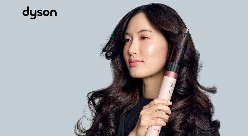 Person mit langen, voluminösen, glänzend gestylten Haaren hält einen Dyson Airwrap Multi-Styler in der Farbe Keramik Pink/Roségold vor hellgrauem Hintergrund; das Dyson Logo ist oben links zu sehen.