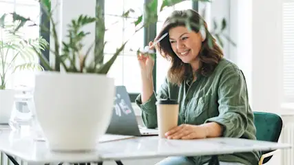 Lächelnde Person in grünem Hemd am Laptop mit Kaffeetasse und Stift. Weiße Töpfe mit Pflanzen im Vordergrund, Fenster im Hintergrund.