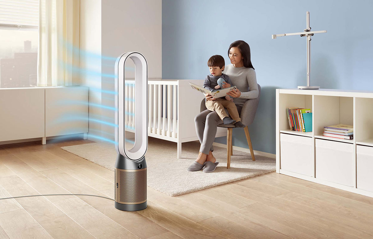 Frau liest Kind ein Buch vor. Im Vordergrund Ventilator mit blauen Luftströmen. Im Hintergrund Gitterbett und Regal.