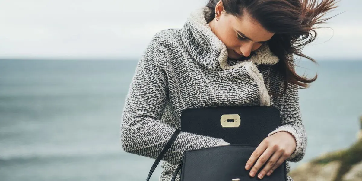 Frau in dicker Strickjacke steht am Meer und öffnet eine schwarze Handtasche.