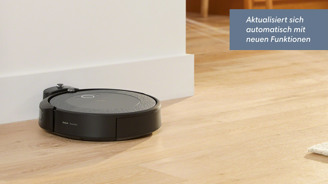 Ein runder, schwarzer Staubsaugerroboter steht auf einem hellen Holzboden neben einer weißen Wand.
