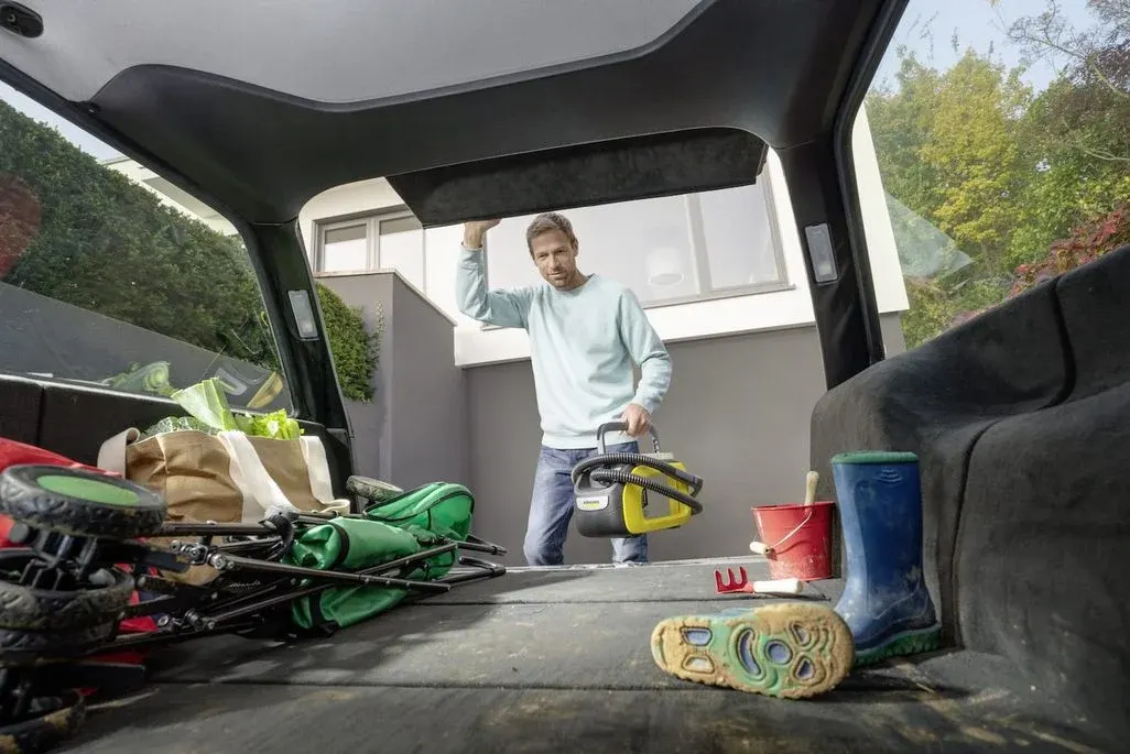 Ein Mann im hellblauen Pullover lädt einen gelb-schwarzen Hochdruckreiniger in den Kofferraum. Dort liegen Einkaufstüten, ein grüner Bollerwagen, rote Gartengeräte und blaue Gummistiefel.