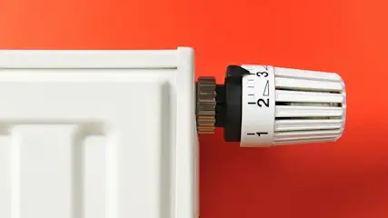 Ein weißer Heizkörper mit einem weißen Thermostatventil, eingestellt zwischen 2 und 3, ist vor einer roten Wand zu sehen.