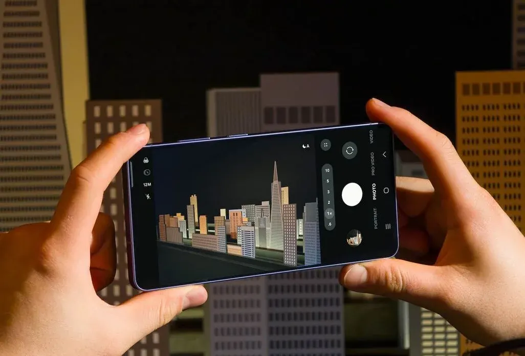 Zwei Hände halten ein Smartphone im Querformat, auf dessen Kameradisplay eine beleuchtete Skyline aus Modellhochhäusern zu sehen ist.