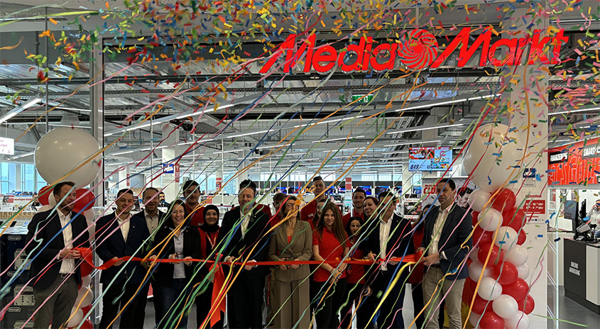 Eine Gruppe schneidet ein rotes Band unter einem MediaMarkt-Schild mit Luftballons und Konfetti.