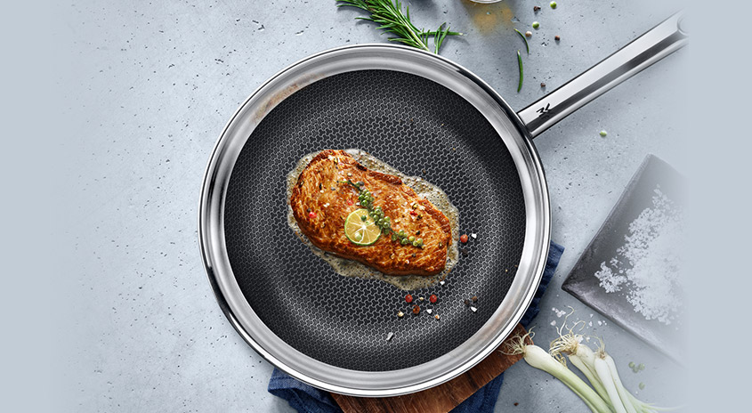 Eine Bratpfanne mit silbernem Rand und Griff. Darin ein Steak garniert mit Limette und Gewürzen. Zwiebeln und Rosmarin liegen daneben.