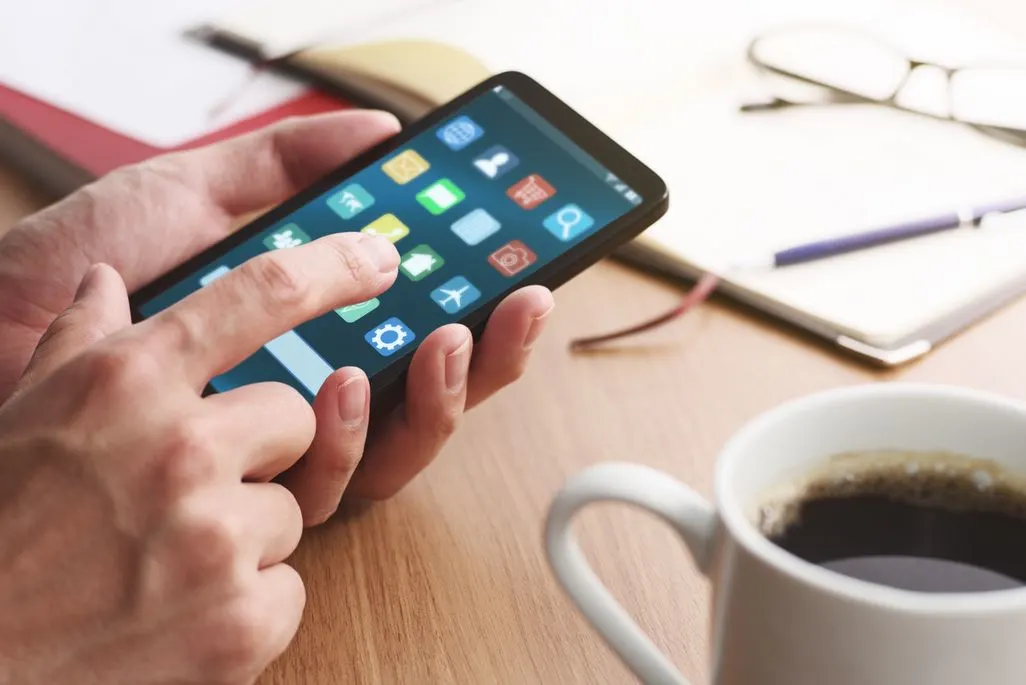 Hand tippt auf App-Symbol auf Smartphone neben einer Tasse Kaffee.