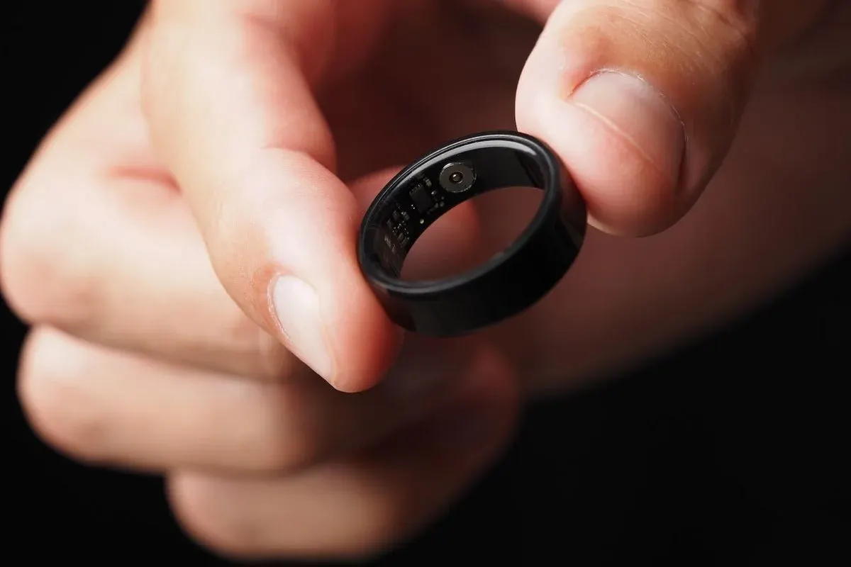 Nahaufnahme einer Hand, die einen schwarzen elektronischen Ring (Smart-Ring) zwischen Daumen und Zeigefinger hält; die Innenseite des Rings mit kleinen Sensoren und Schaltkreisen ist sichtbar.