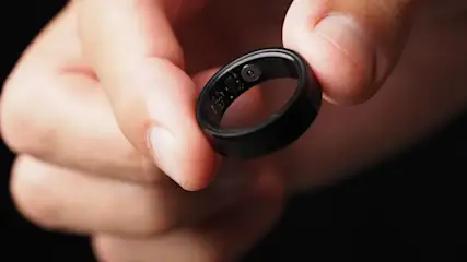 Nahaufnahme einer Hand, die einen schwarzen elektronischen Ring (Smart-Ring) zwischen Daumen und Zeigefinger hält; die Innenseite des Rings mit kleinen Sensoren und Schaltkreisen ist sichtbar.