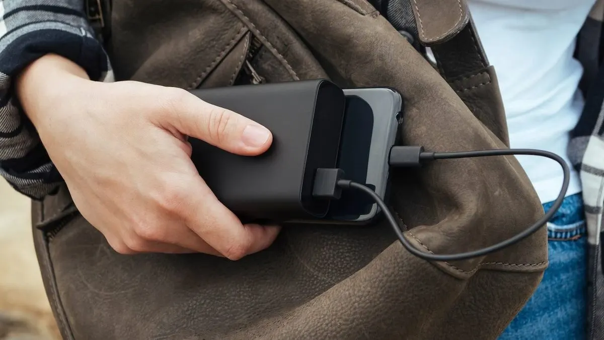 Hand steckt eine Powerbank und ein Smartphone in eine braune Umhängetasche, während das Handy über ein Kabel geladen wird.