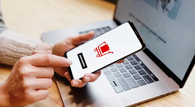 Eine Person hält ein Smartphone mit rotem Einkaufswagen-Symbol und "Jetzt kaufen"-Button über einem Laptop.