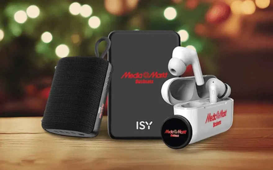 On a wooden table are electronic gadgets with the MediaMarkt logo: a speaker, a powerbank and wireless earbuds, set against festive lights.