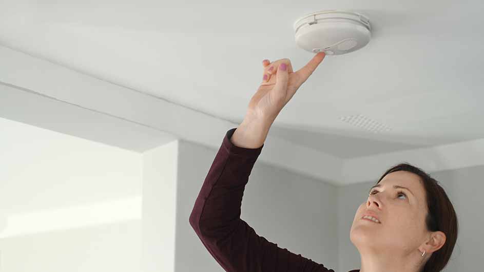 Une main appuie sur un détecteur de fumée blanc fixé au plafond blanc d'une pièce.