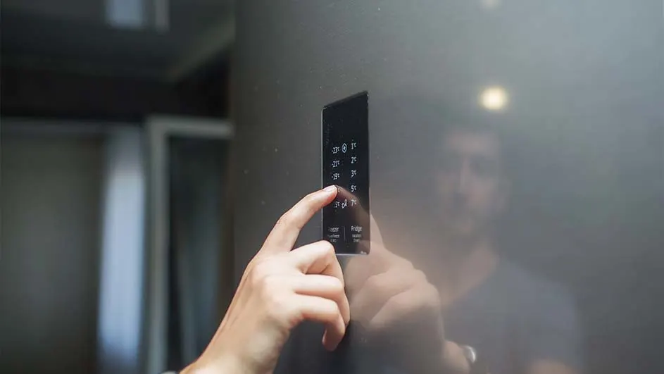 Une main touche le panneau de commande noir d’un réfrigérateur moderne. Sa surface brillante reflète la personne et la cuisine sombre située à l’arrière-plan.
