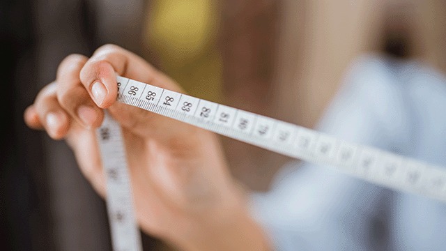 Een witte meetlint met zwarte cijfers wordt vastgehouden door een hand, tegen een vage achtergrond.