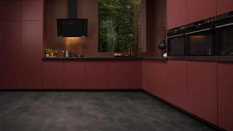 Een keuken met bordeauxrode kasten, een zwart aanrecht en zwarte apparaten. Buiten is er een bos zichtbaar.