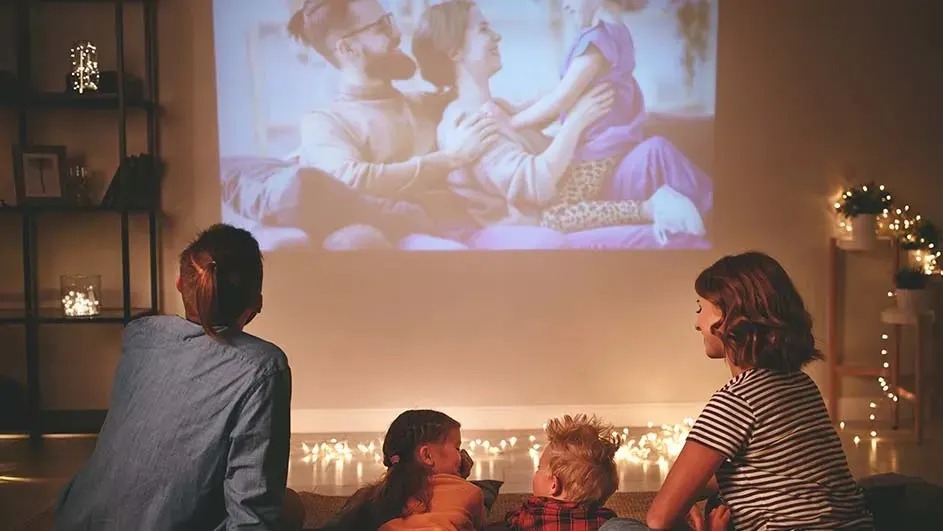 Une famille est assise confortablement sur le sol d’un salon chaleureux et regarde un film projeté sur le mur. Aucune marque n’est visible.
