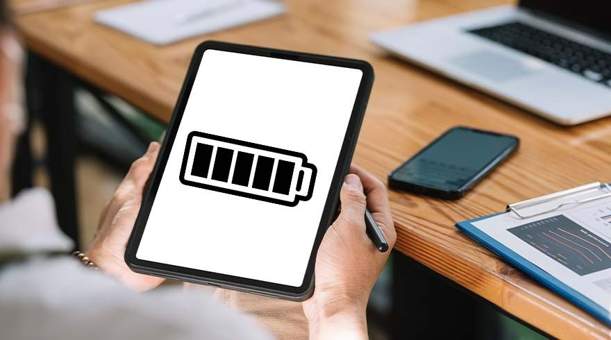 Een persoon houdt een tablet vast met een batterijpictogram. Een telefoon, laptop en klembord liggen op een houten bureau.