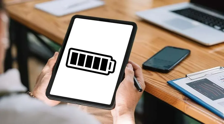 Een persoon houdt een tablet vast met een batterijpictogram. Een telefoon, laptop en klembord liggen op een houten bureau.
