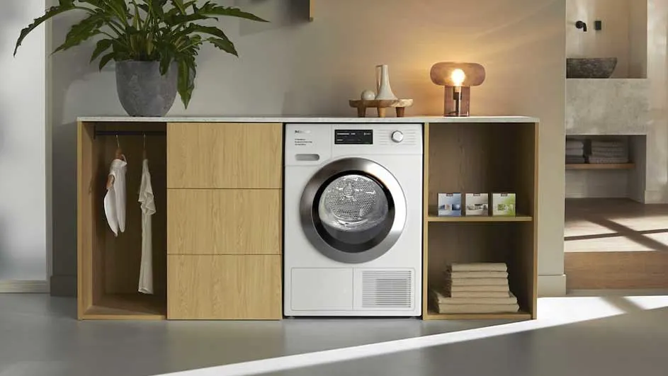 Een kamer met een witte droogkast tussen houten kasten, een plant erboven en een lamp. Op de achtergrond is een wastafel te zien.